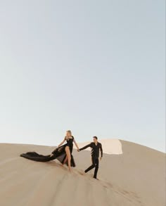 a man and woman in black clothes are walking across the sand dunes with their arms around each other