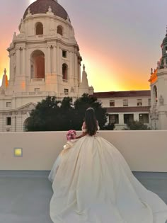 a woman in a wedding dress is sitting on a ledge looking at the sun set