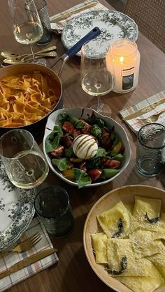 a wooden table topped with plates and bowls filled with food next to glasses of wine