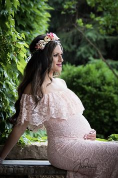 a pregnant woman in a pink dress sitting on a bench with greenery behind her