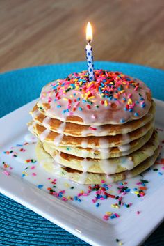 a stack of pancakes on a plate with a lit candle in the middle and sprinkles all around it