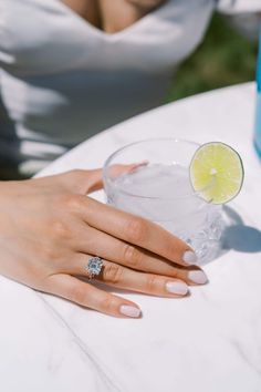 a woman sitting at a table holding a glass with a lemon slice on it and a diamond ring