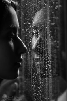 black and white photograph of woman looking out window with raindrops on it's glass