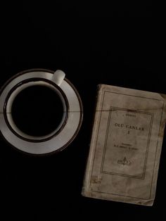 an old book next to a coffee cup and saucer on a black table top