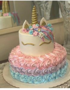 a three layer cake decorated with pink, blue and white frosting has a unicorn's face on top