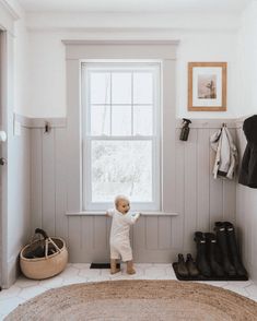 a toddler standing in front of a window with shoes on the floor next to it