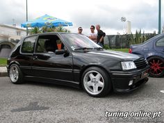 two men standing next to a black car in a parking lot with other cars behind them