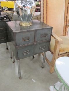 an old metal cabinet with a silver vase on top and two chairs in the background