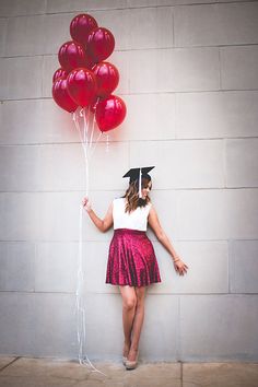 a woman in a graduation cap and gown holding red balloons while standing against a wall