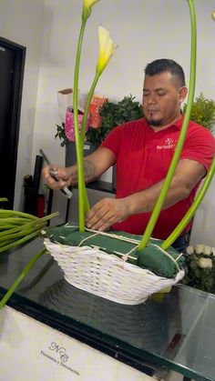 a man cutting up flowers in a basket on a glass table with other plants behind him