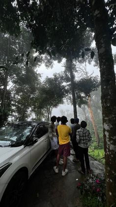 several people standing next to a car on a road in the rain with trees around it