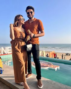 a man and woman standing next to each other near the ocean with people in the background