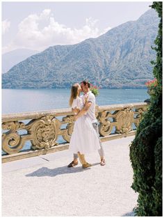 a man and woman are kissing on a bridge by the water with mountains in the background