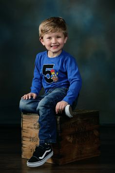 a young boy sitting on top of a wooden crate smiling at the camera while wearing a blue shirt and jeans