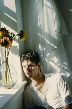 a man sitting in front of a window next to a vase with sunflowers