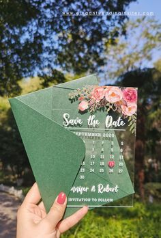 a person holding up a green save the date card with pink flowers on it and greenery in the background