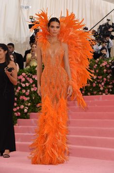 a woman in an orange feathered dress stands on the pink carpet with cameras around her