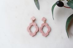 two pink earrings sitting on top of a white table next to green leaves and a potted plant