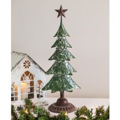 a small christmas tree next to a lit up house on a white table with greenery