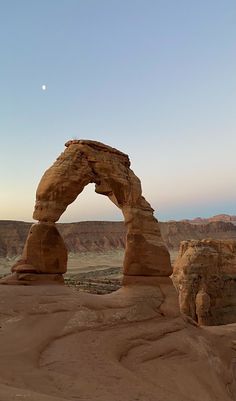 an arch shaped rock formation in the middle of desert with a moon visible above it