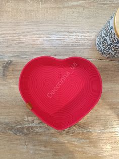 a red heart shaped basket sitting on top of a wooden floor next to a vase