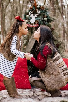 two women sitting on the ground in front of a christmas tree with snow falling all around them