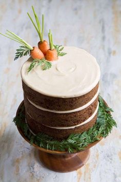 a cake with carrots on top is sitting on a wooden stand and has greenery around it