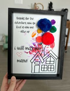 a hand holding up a framed artwork with words written on it and pom - poms