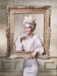 a woman with white hair is posing in front of a gold framed painting and holding her hand on her chin