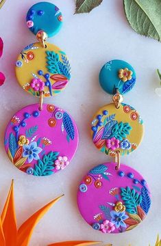 three colorful earrings sitting on top of a white surface next to flowers and green leaves