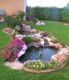 a small pond in the middle of a lawn with flowers and rocks around it, next to a house