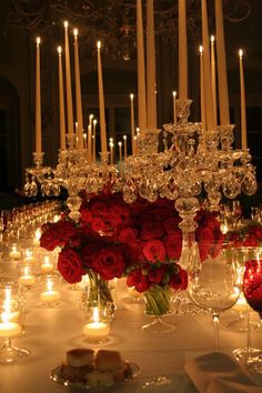 a table with candles and red roses in vases on it, surrounded by wine glasses