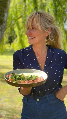 a woman holding a plate with food on it in the grass and trees behind her