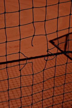 the shadow of a tennis racket is cast on an orange clay court with black netting