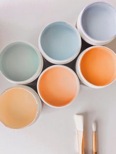 five different colors of paint in white bowls next to two brushes and one tube of toothpaste