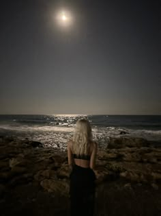 a woman standing on top of a rocky beach next to the ocean under a full moon