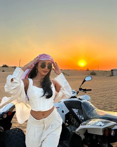 a woman standing next to a motorcycle in the desert at sunset or sunrise, wearing sunglasses and a head scarf