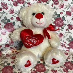 a white teddy bear with a red heart on it's chest sitting in front of a floral background