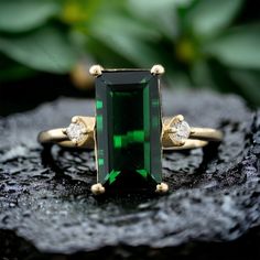 an emerald and diamond ring sitting on top of a piece of rock with leaves in the background