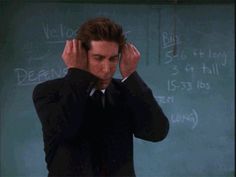 a man in a suit and tie holding his head to his ears while standing in front of a chalkboard