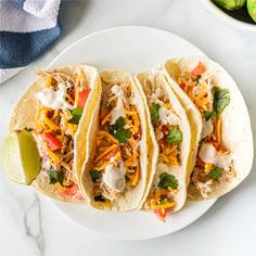 three shredded chicken tacos on a white plate with limes and cilantro