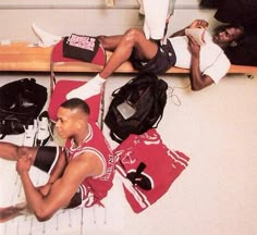 two young men sitting on the floor with backpacks and sports gear in front of them