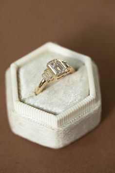 a three stone diamond ring sits in a white box on top of a brown surface