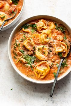 two bowls filled with pasta and sauce on top of a white table next to silver spoons