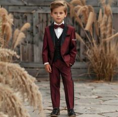 a young boy in a red suit and bow tie standing on a brick walkway next to tall grass