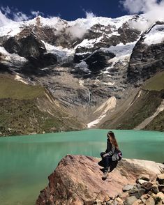 a woman sitting on top of a large rock next to a lake with snow covered mountains in the background