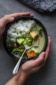 two hands holding a bowl filled with rice, broccoli and tofu soup