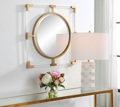 a vase with flowers on a table next to a round mirror and lamp in a white room