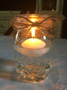 a lit candle in a glass jar on a white table cloth with a bow around it