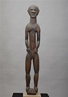 an old wooden statue is standing on a black platform with a gray background and white wall behind it